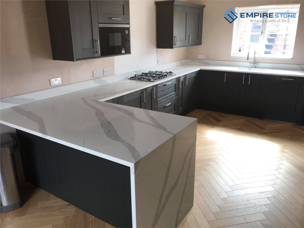 Bếp chữ U tủ đen mặt đá trắng vân xám, đầu bán đảo làm kiểu thác nước trên sàn gỗ xương cá