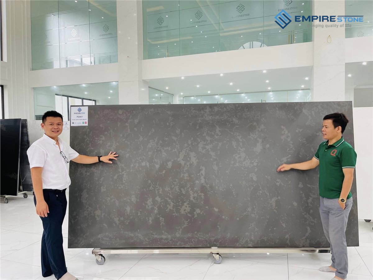 Đá nhân tạo cho văn phòng: Nên dùng ở đâu và chọn màu thế nào cho chuyên nghiệp? Hai người đứng cạnh tấm đá nhân tạo màu xám đậm khổ lớn đặt trên giá trong showroom
